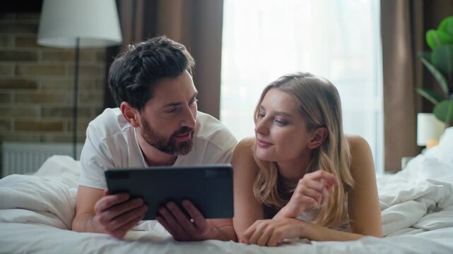 Concentrated couple looking in digital tablet screen display talking smile laugh in bed at morning. Caucasian family man with woman talk use portable computer smiling laughing affectionate bonding