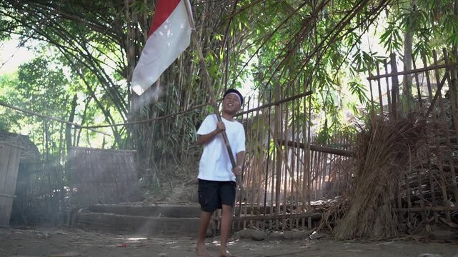 Person walks forward beside tall red and white flag in dusty rural village path, with bamboo fence, tropical trees, bright daylight, soft haze, and rustic steps, creating calm local documentary mood