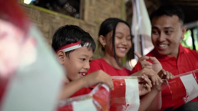 Asian family gathers indoors in warm festive light, holding red and white flag bunting while child stands in foreground and smiling adults pose behind rustic woven wall, with family, child, adult
