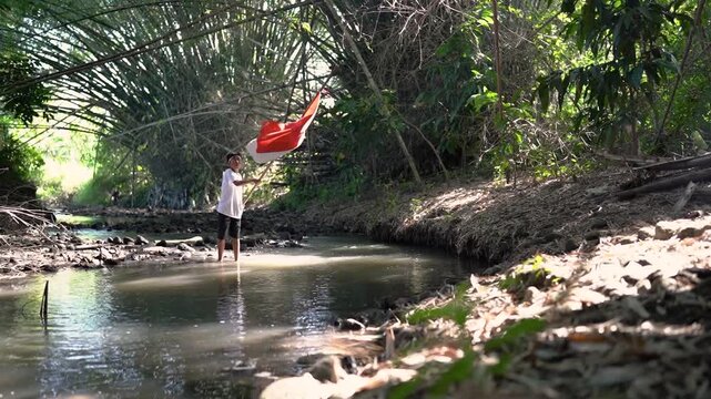 Young boy stands barefoot in shallow creek and raises red and white flag on tall pole beside dense tropical vegetation, with rocky banks, tangled branches, reflective water, and soft sunlight