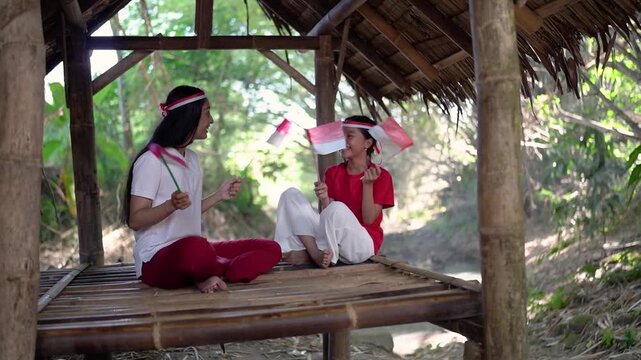 Young girls sit cross legged on bamboo platform inside rustic thatched shelter, smiling and facing each other while holding small red and white flags amid tropical greenery and daylight