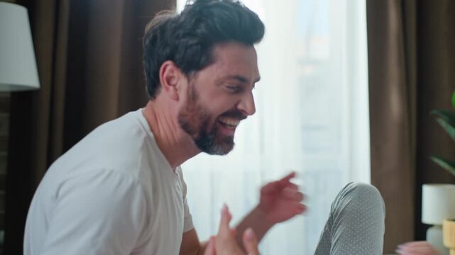 Happy Caucasian family married couple fooling around having fun tickling laugh in bedroom awaking in morning together. Playful husband man tickle wife woman in bed laughing candid romantic honeymoon