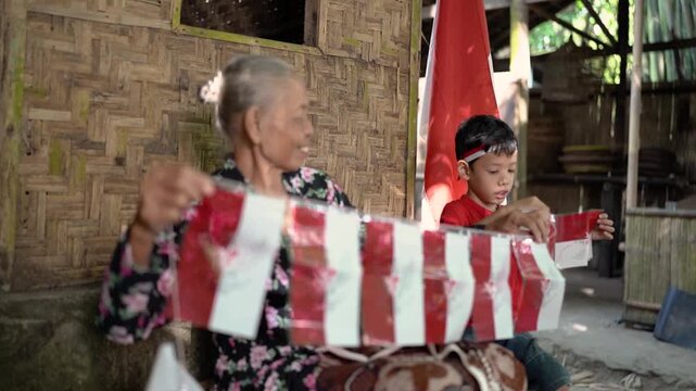 Smiling older woman and young boy sit beside woven bamboo wall in rustic village home, actively handling red and white flag ribbons with blurred hands and foreground fabric, creating cheerful