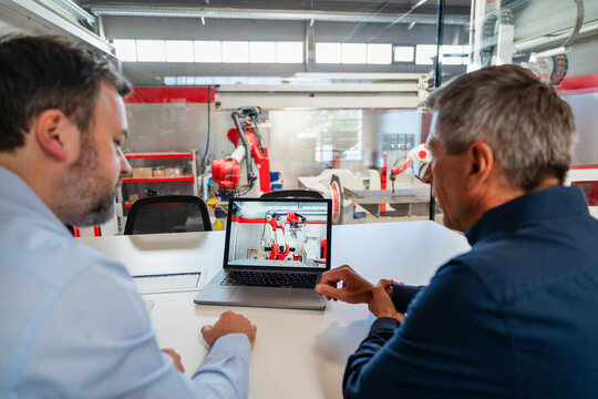 Two engineers monitor an industrial robot on a laptop in a factory