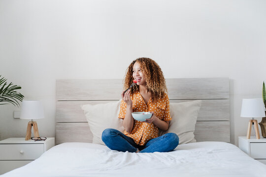 Young woman eating fruit salad while sitting with crossed legs on bed