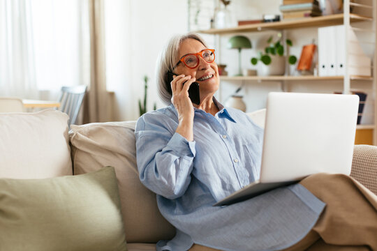 Smiling woman talking on smart phone sitting with laptop in living room