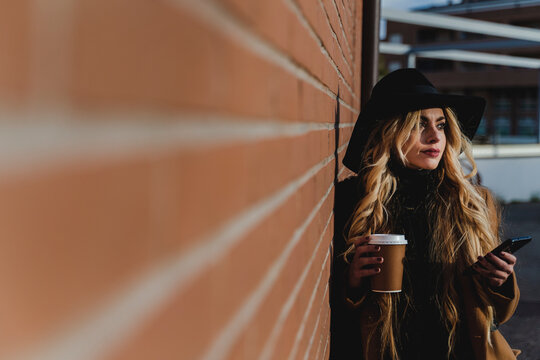 Young woman in hat with coffee cup standing by brick wall while looking away