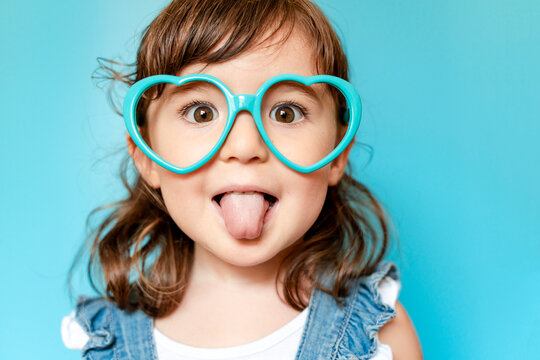 Portrait of cute little girl with tongue out and heart shaped glasses on blue background