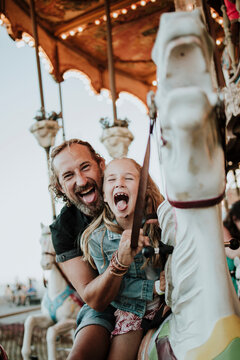 Father and daughter sticking out tongue enjoying carousel ride at amusement park