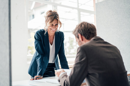 Happy businesswoman leaning on desk looking at client in office