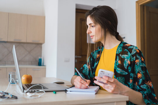 Young woman sitting at table at home using cell phone and laptop taking notes