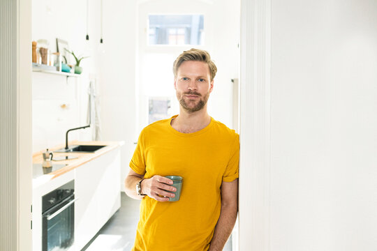 Confident casual man with coffee mug leaning against door case