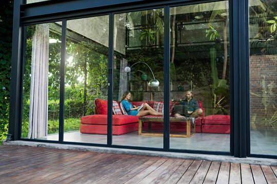 Garden view through glass facade of couple with laptop and book relaxing on red couch in modern design house