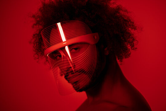Close-up of young man wearing red led light mask against wall at home