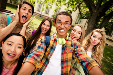 Fototapeta premium Group of happy diverse college friends taking a selfie on a leafy campus, smiling students with backpacks and headphones enjoying outdoor time together