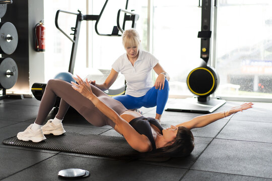 Female trainer guiding woman through supervised floor rehabilitation exercise in bright gym with mat work, mobility focus, core strengthening and supportive coaching