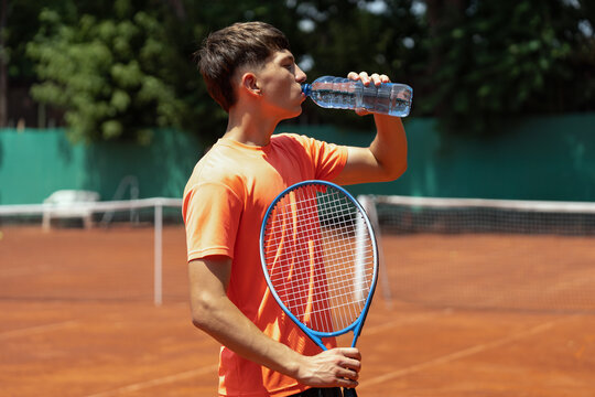 Teenage tennis player drinking water while holding racket during break on an outdoor clay court in bright summer sunlight