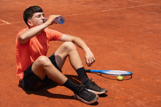 Junior tennis player rehydrating with water on clay court after intensive training or competition on hot sunny day. Summer sports, recovery, hydration and healthy lifestyle concept
