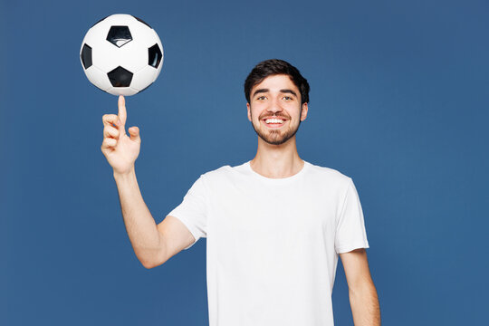 Young smiling happy cheerful fun man fan wear basic white t-shirt cheer up support football sport team hold in hand spin soccer ball watch tv live stream look camera isolated on plain blue background.