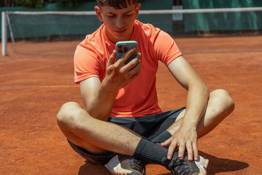 Teenage tennis player using smartphone while sitting on clay court during break between training sessions, combining sports, mobile apps, sports technology, communication and recovery