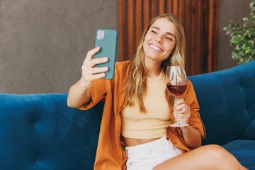 Young woman wear casual clothes sits on blue sofa couch do selfie shot on mobile cell phone drink wine stay at home hotel flat rest relax spend free spare time in living room indoor. Lounge concept. © ViDi Studio
