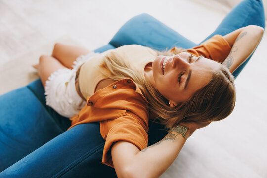 Young minded calm woman wearing casual clothes hold hands behind neck sit on blue sofa stay at home hotel flat rest relax spend free spare time in living room indoors grey wall. People lounge concept.