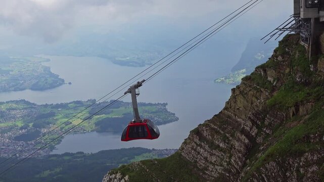 Red cable car descends from Mount Pilatus above Lake Lucerne in Switzerland, with rocky cliffs, misty Alps, and clouds creating a dramatic mountain travel scene. 4K UHD video.