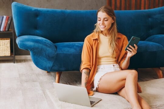 Young woman wear brown shirt use work on laptop pc computer hold mobile phone sit at blue sofa couch stay at home hotel flat relax spend free spare time in living room indoors. People lounge concept.