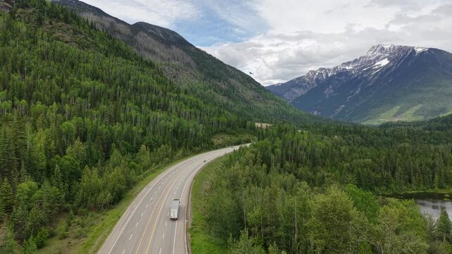 Aerial drone footage of a pristine mountain valley with a reflective pond dense pine forest a winding road and snowcapped peaks in the distance along the Canadian Rockies in Revelstoke, BC.