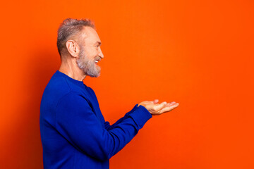 Senior man smiling and presenting with open palms in blue sweater against bright orange background, cheerful profile portrait and copy space for advertising or lifestyle concept © deagreez