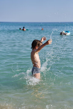 A cheerful child enjoys playing in the refreshing turquoise ocean waves, throwing water with arms raised during a family beach holiday.