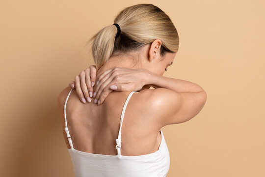 Young beautiful woman massaging her neck and upper back on beige studio background, representing muscle tension, body pain relief and self care wellness