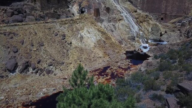 Scenic view looking into the valley of the Rio Tinto river featuring a beautiful small waterfall with flowing, mineral-rich water. It showcases a historic dam wall and an old, abandoned stone building