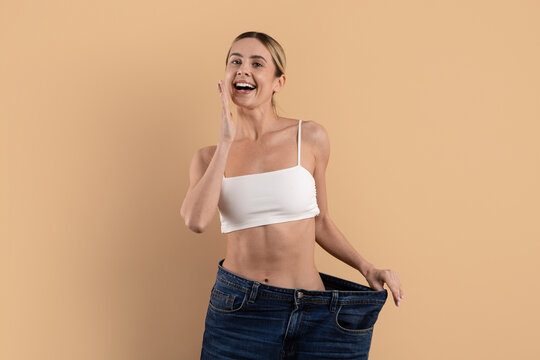 Young beautiful woman laughing and showing oversized jeans on beige studio background, expressing successful weight loss, confidence and healthy lifestyle