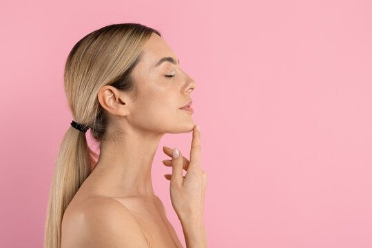 Young beautiful woman in side profile touching her chin with eyes closed, posing on pink studio background for skincare, facial care and beauty concepts, copy space