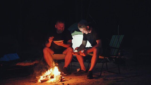 Two men sit inside tent beside campfire at night, sharing quiet meal and conversation in warm firelight, with folding chair, glowing tent opening, dark forest camp setting, rustic outdoor atmosphere