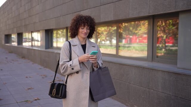A stylish young woman checks her smartphone outdoors in a vibrant city while wearing a coat