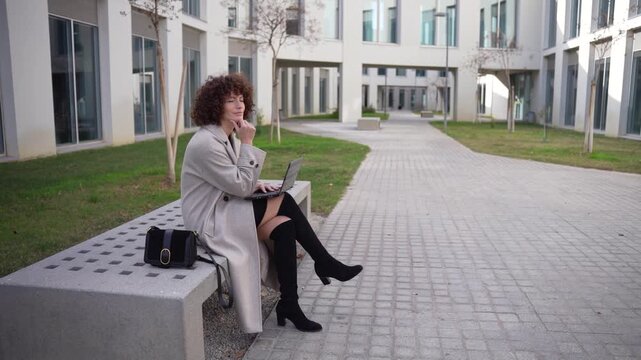 A woman is working diligently outdoors on her laptop within a modern urban environment