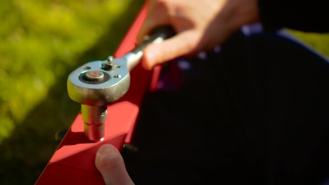 Close up ratchet wrench tightening bolt on red steel bracket outdoors. Precision fastening process with hand tool and metal hardware in sunlight. Engineering assembly detail with mechanic focusing on