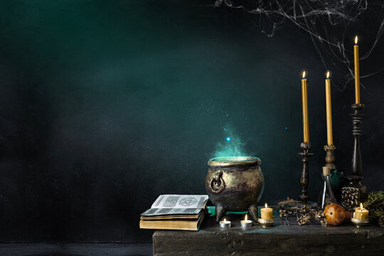 Halloween background. Dark witch cauldron with a magic witch's potion, books, spell book. Witchcraft paraphernalia and accessories among candles on a table in a room or cave with stone walls.