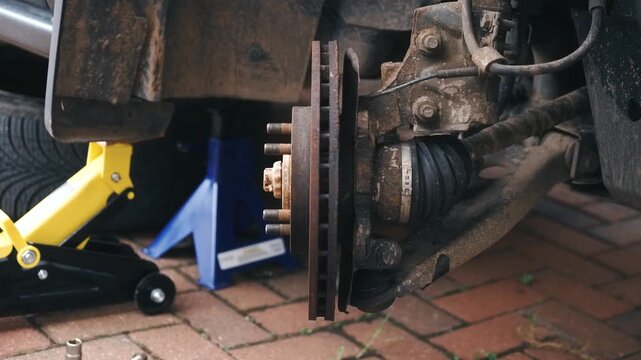 Raised car with removed front wheel, rusty brake disc and worn brake pads, exposed suspension components. Automotive maintenance concept with hydraulic jack, wheel hub, axle assembly on driveway