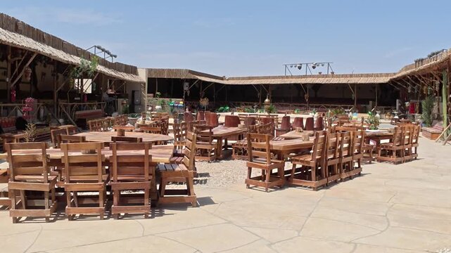 Cinematic view of a spacious open air restaurant at a traditional desert camp featuring handcrafted wooden tables and chairs under rustic shaded structures. Authentic Middle Eastern style.