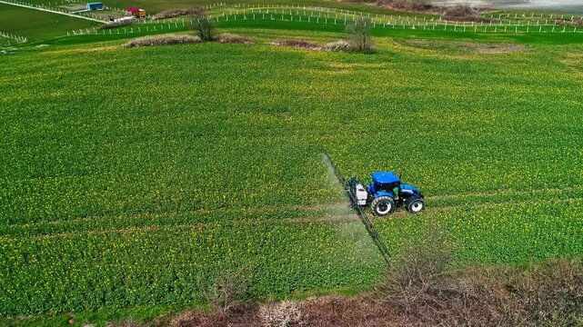 A drone view shows a tractor-mounted sprayer working on green farmland, performing agricultural spraying for crop protection, field maintenance, and modern farming practice.