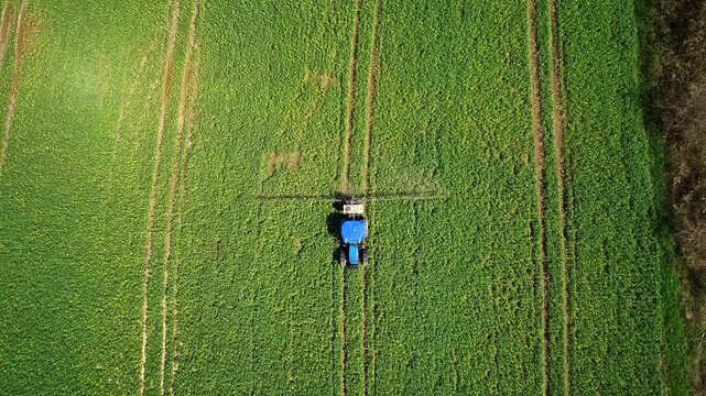 A drone view shows a tractor-mounted sprayer working on green farmland, performing agricultural spraying for crop protection, field maintenance, and modern farming practice.