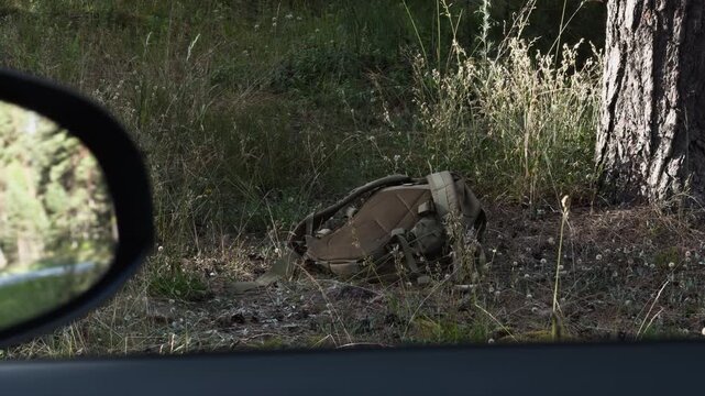 Abandoned hiking backpack lying beside tree in grassy forest clearing seen from parked vehicle