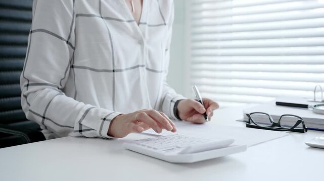 Businesswoman hands calculating finance, bookkeeping, and taxes, using an office calculator while writing down important financial data on documents at a desk