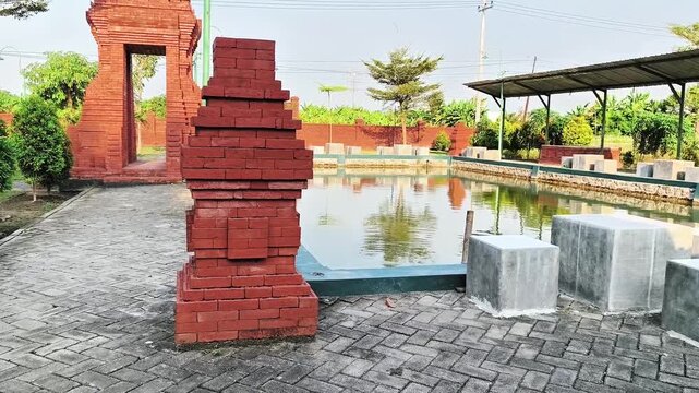 Miniature Majapahit style red brick temple gate or gapura by a scenic fish pond in a cultural park resort, traditional Javanese heritage architecture in Indonesia.