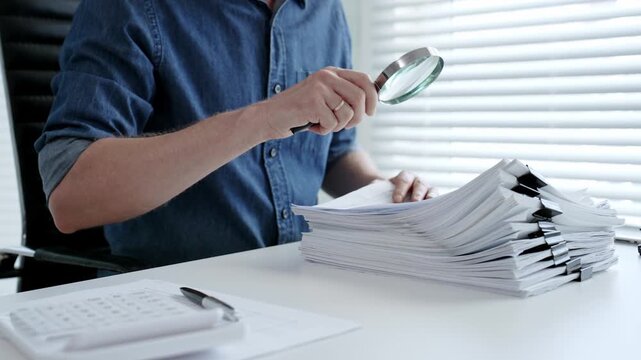Person examining business documents with a magnifying glass, performing a detailed audit, financial analysis, legal investigation, and careful review of paperwork for accuracy