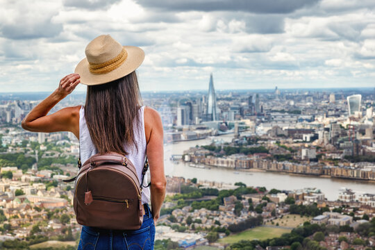 A tourist woman with a backpack looks at the elevated view of the skyline of London, England, during a sunny summer day