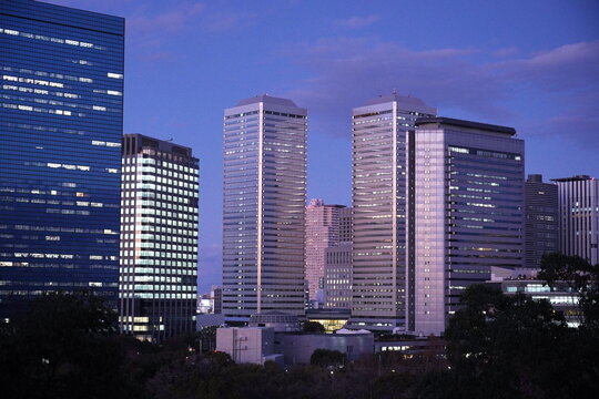 大阪の夕暮れ時、多くの窓に明かりが灯る高層ビル群が織りなす現代的で美しいビジネス街の夜景。